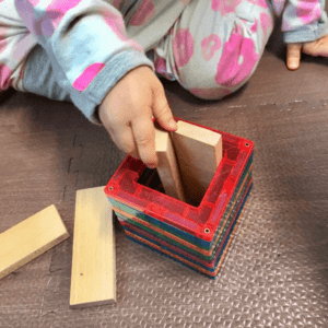  A baby in the posting schema, carefully placing a rectangular wooden block inside her magnetic tile structure. 