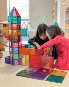 Siblings collaborate to construct story-inspired structures like castles and buildings using magnetic tiles. They enthusiastically share ideas and arrange the tiles, fostering teamwork and creativity. 