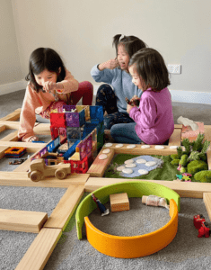 Siblings immersed in imaginative play, using magnetic tiles to build houses, trees, and a farm shelter, demonstrating their ability to recreate real-world environments and expand their storytelling skills through open-ended play. 