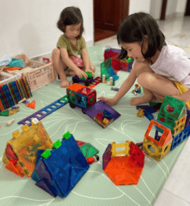 Siblings are playing together to create a small world using colourrful magnetic tiles. There are construction vehicles, road, and houses of different designs. 