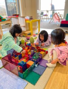 Siblings are immersed in imaginative play, using magnetic tiles to build a house-like structure and created a small world setting. As they construct their scene, they share stories and engage in creative dialogue, demonstrating their ability to recreate real-world environments and enhance their storytelling skills through open-ended play. 