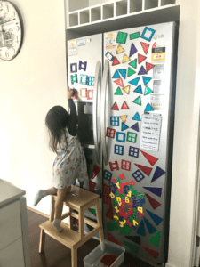 A toddler carefully sticks the magnetic tiles to the fridge door, grouping different shapes together. The fridge door is a vertical surface and allow the child to experiment with balance, gravity, and spatial orientation. 