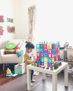 Toddler is constructing a castle using magnetic tiles, surrounded by loose parts like coins, rings, and cups. Peg dolls are placed around the castle-like structure, enhancing the imaginative play 