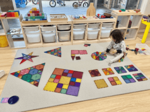 Shapes creation using magnetic tiles, combining hexagons and triangles to form a larger triangle, while also using various sizes of squares to construct a big square. A toddler is placing rainbow acrylic blocks on the magnetic tiles dodecagon corresponding to its colour. 