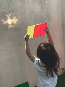 A toddler using magnetic tiles to explore light and reflections, experimenting with how sunlight passes through transparent tiles, fostering curiosity about science and visual effects.