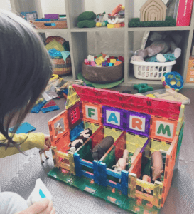 A toddler is creating a farm shelter using colourful magnetic tiles and arranging the tiles to create separate areas for various animal figurines placed inside the structure.