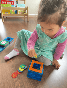 A young toddler carefully places a coin inside her magnetic tile structure, adding to the creative build. The coins are numbered, and the toddler posts each one thoughtfully, practicing number recognition and early counting skills as part of her imaginative play. 