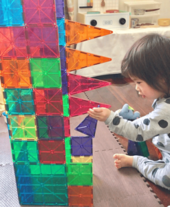 A toddler places triangle magnetic tiles sideways on a tall tower structure, exploring how the magnets hold. 