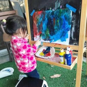 A toddler painting in a cardboard using blue, red and yellow paints