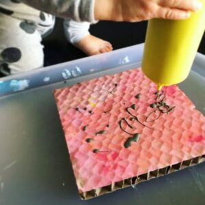 Toddler squeezing a bottle of coloured glue on an existing artwork on a corrugated cardboard canvas.