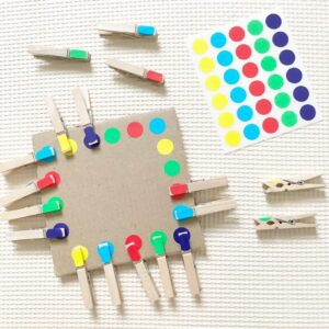 A toddler activity setup using recyclable cardboard, wooden pegs, and colorful dot stickers. Toddler match the colour on the pegs to the dot stickers on the corrugated cardboard.