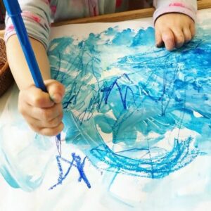 Toddler using blue markers to draw additional details on a previously created piece of artwork.