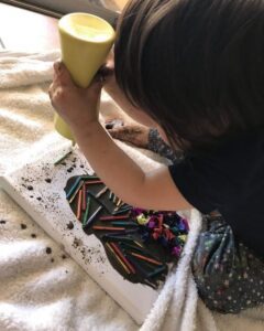 A toddler is creating a collage on an existing black canvas, carefully placing various colorful materials onto the dark background.