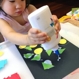 Toddler squeezing liquid glue onto a black sheet of paper and sticking various shapes cut from colorful construction paper onto the glue. The child’s focused hands are actively engaged in creating a vibrant collage with the bright shapes and glue.