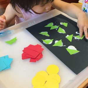 Toddler creating a collage by arranging various colourful shapes cut from construction paper and sticking them onto a black sheet of paper.