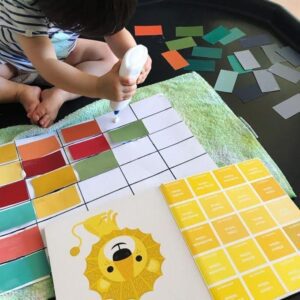 Toddler carefully sticking colorful paint chips onto a grid canvas, creating a vibrant and patterned artwork. Pantone colour book to show toddler the different hues of yellow. 