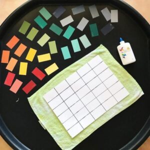 Paint chips and grid paper arranged as materials for a toddler process art invitation. The paint chips display a range of colors, and the grid paper provides a structured backdrop for creative exploration and art activities.