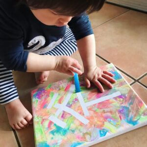 Toddler carefully peeling washi tape from a canvas after a painting session, revealing a clean negative space where the tape was placed. The surrounding canvas is covered in colorful paint, highlighting the contrast created by the removed tape.