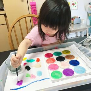 Toddler painting circles with a brush and watercolor palette, carefully dipping the brush into vibrant colors and applying them to paper. The child's focused expression and steady hand capture the joy of exploring art through playful, colorful strokes.