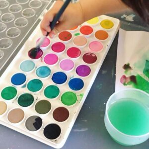 An inviting watercolor painting setup for a toddler, featuring a watercolor palette with a range of bright colors, a small brush, a cup of water, and a sheet of white paper. Toddler picking up paints from the watercolour palette. 