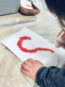 Toddler carefully stamping a red dot marker to form the letter 'S' on a white piece of paper. The child’s small hand grips the marker with focus, creating a vibrant and playful design as each dot takes shape in the curve of the letter.