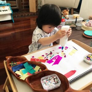 Toddler squirting glue onto bits of paper, googly eyes, and craft pieces to create a collage. The child’s hands are actively applying glue and arranging the materials on a surface, focusing on assembling a colorful and textured artwork.