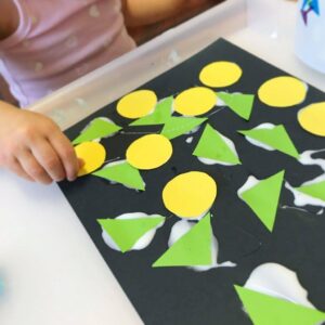 Toddler creating a collage by arranging various colourful shapes cut from construction paper and sticking them onto a black sheet of paper.