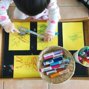 Toddler scribbling on a yellow piece of paper with washable markers. A basket filled with an assortment of washable markers set up as an invitation to create. 