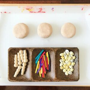 Invitation to create with playdough and loose parts, including popsicle sticks, shells, and small pieces of play dough. The materials are arranged on a surface, encouraging imaginative play and hands-on creativity with a variety of textures and shapes.