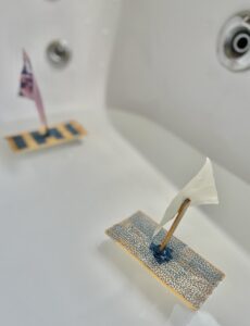 Two homemade boat crafted from household items, being tested for floatability in a bathtub. The boat is floating on the water as the child observes its stability and buoyancy.