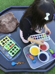 A toddler exploring colour mixing with coloured water using various tools such as droppers, ice tray and small cups, The child is experimenting and creating new colours.