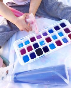 A toddler using a spray bottle filled with red and blue primary colors to create different shades of purple and blue on an ice tray. The child is experimenting with color mixing and spray techniques, creating vibrant hues on the ice.
