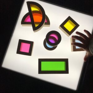 A toddler arranging colourful rainbow blocks on a light table, with the soft glow of the table illuminating the blocks, creating a vibrant display of colours. 
