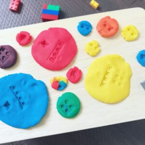 Three primary coloured play dough (red, blue, and yellow) being mixed to create secondary colours (orange, green, and purple) in smaller piles. LEGO pieces are used to make imprints and marks on the play dough, showcasing the creative blending and texture.
