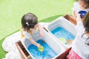 Three siblings happily engaging in water play, using blue-coloured water and containers of different sizes at a water play station. 