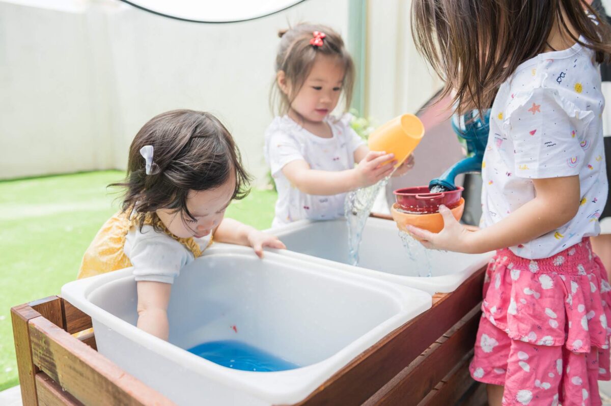 Siblings pouring water using cups, bowls, and a toy watering can, exploring different tools for water play