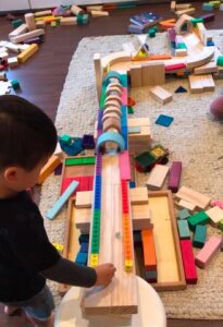 A preschooler building a ball run ramp with blocks, arranging unifix cubes along the tiles as a fence, and using a rainbow stacker as tunnels for the ball to roll through.
