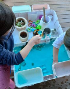 Two children engaging in ice play with household items and natural materials on a Kingdom Play table, exploring textures and observing the melting process of the ice.
